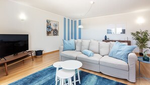 Ein modernes Wohnzimmer mit einer Couch, zwei weißen Beistelltischen, einem Flachbildfernseher, einer blau gestreiften Wand und einer Topfpflanze.