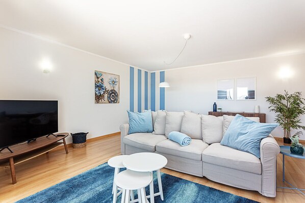 Un salon moderne avec un canapé, deux petites tables d’appoint blanches, un téléviseur à écran plat, un mur à rayures bleues et une plante en pot.