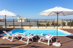 Un espace piscine aménagé sur le toit, avec des chaises longues blanches et des parasols, offrant une vue dégagée sur le paysage urbain en arrière-plan.