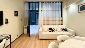 Un dormitorio moderno con sofá cama, una mesa de comedor con sillas, una mesita de noche con lámpara y un amplio ventanal con cortinas translúcidas.