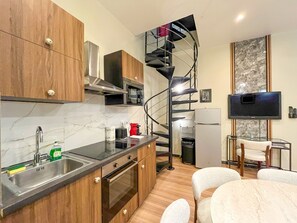 A compact kitchen with wooden cabinets, a stainless steel sink, a microwave, and a built-in oven. A spiral staircase leads to a dining area with a table and chairs. - Urban Flat 21 - Modern Duplex in Montorgueil (París)