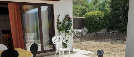 Un bain à remous extérieur surmonté d’une pergola en bois, une porte vitrée donnant sur un espace patio avec des chaises, et une vue sur un jardin et des collines en arrière-plan.