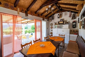 A dining area with wooden tables and benches, a kitchen in the background, and a view of the outdoors through large windows. - Apartment Laurel Leaf (Konavle)