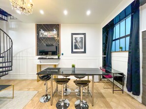 Una cocina moderna con encimera negra, tres taburetes de bar, una lámpara de araña, una escalera y un ventanal con cortinas.
