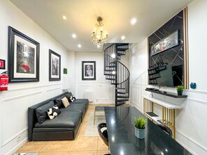 A modern living room with a black sofa, a chandelier, framed pictures on the wall, a staircase, and a TV on the wall.