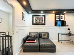 A modern living room with a black leather sofa, patterned cushions, a framed cityscape picture, a small table with a lamp, and a TV on a stand.