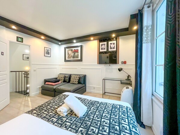 A hotel room with a bed, a sofa, a small table, a mirror, and a window with curtains.