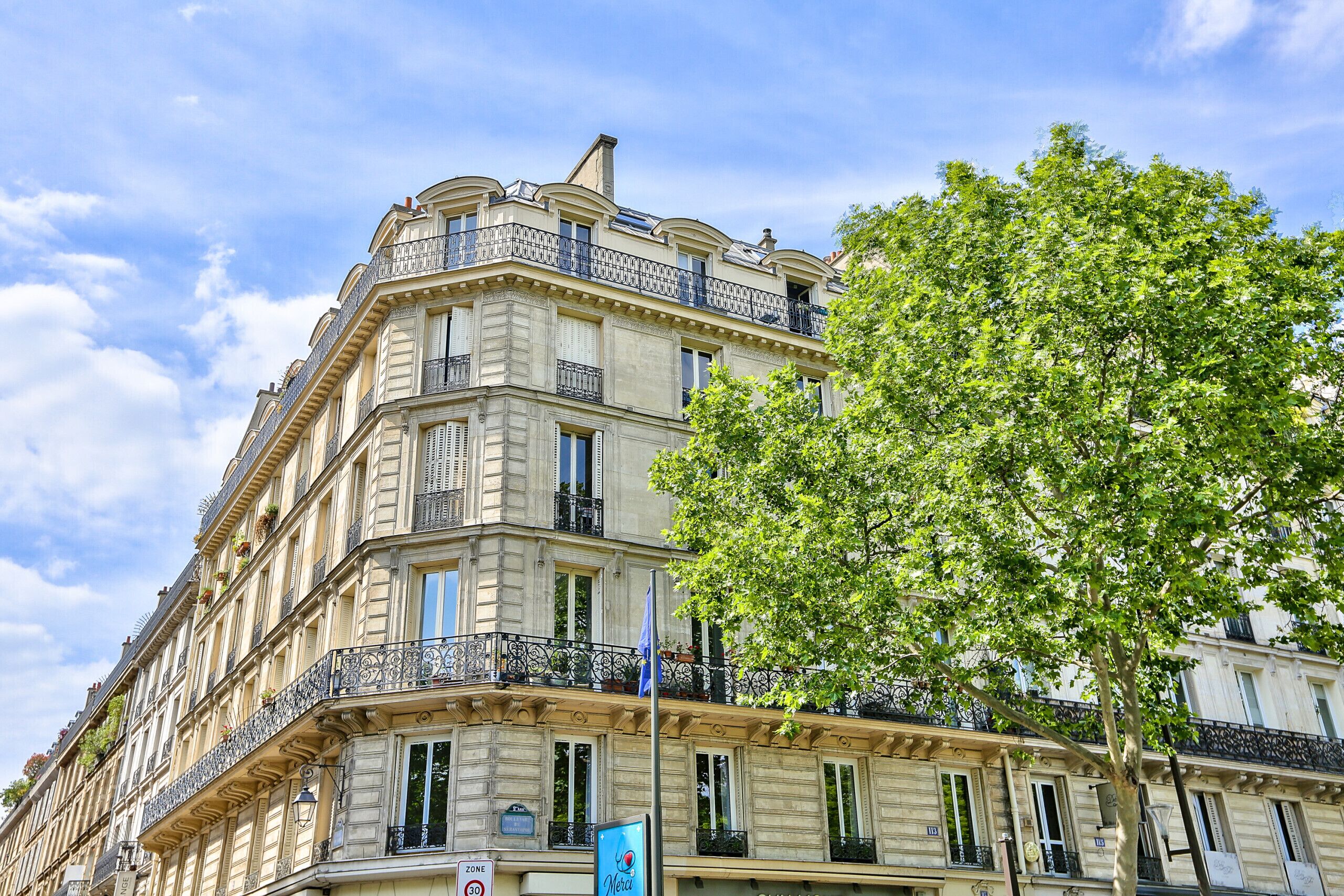 A multi-story building with balconies, a tree in the foreground, and a clear sky.