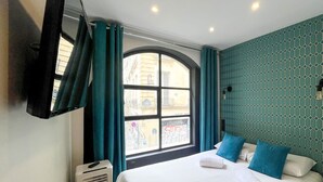 A hotel room with a bed, a window with curtains, a wall with a geometric pattern, and a ceiling with recessed lighting.