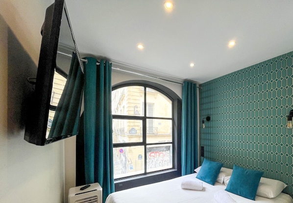 A hotel room with a bed, a window with curtains, a wall with a geometric pattern, and a ceiling with recessed lighting.