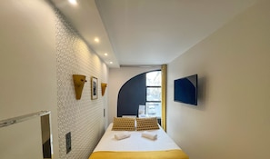A modern hotel room with a patterned wall, a bed with two pillows, a flat-screen TV, and a window with a view of the street.