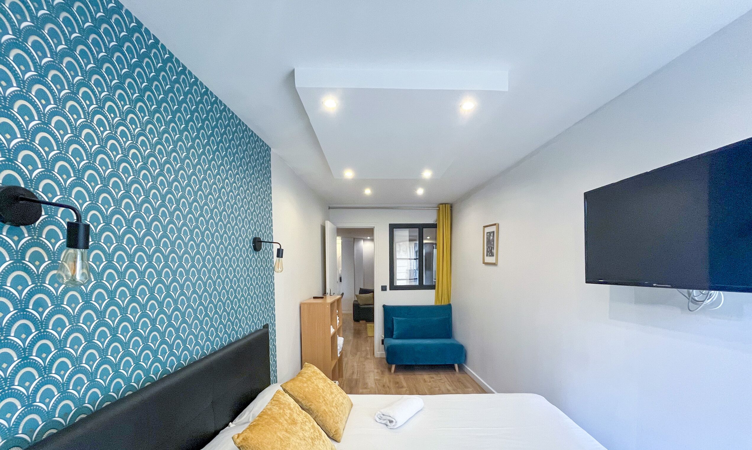 A modern hotel room with a patterned wall, a bed with pillows, a desk, a chair, and a TV mounted on the wall.