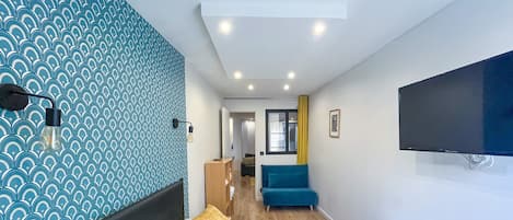 A modern hotel room with a patterned wall, a bed with pillows, a desk, a chair, and a TV mounted on the wall.