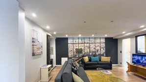 A modern living room with a black sofa, a coffee table, and a TV. There is a large window with a cityscape view.