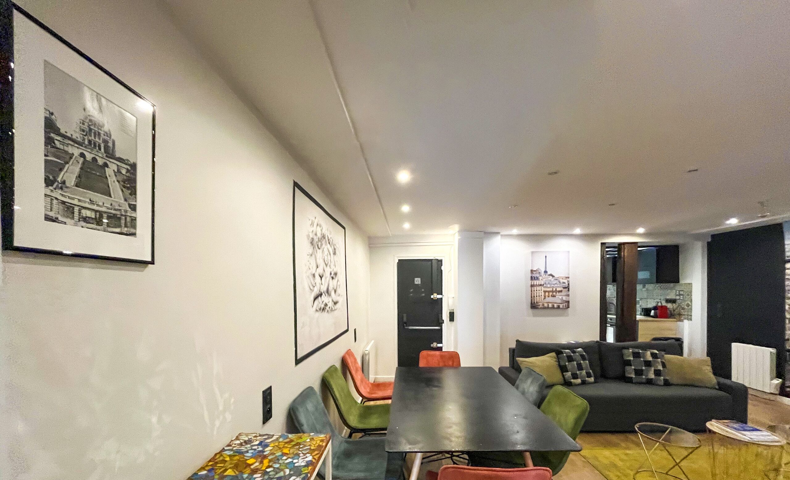A modern living room with a dining area, featuring a sofa, a table, and chairs. There are framed pictures on the walls and a kitchen area in the background.
