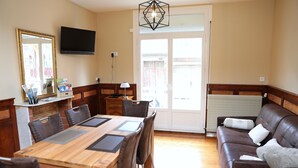 Een eethoek met een houten tafel en stoelen, een bank, een televisie en een groot raam met uitzicht op een gebouw.