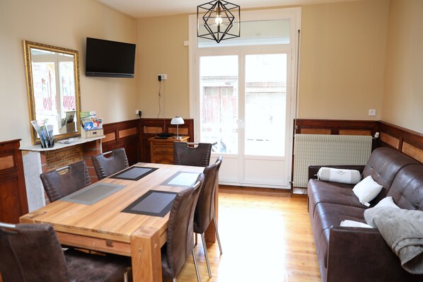 A dining area with a wooden table and chairs, a sofa, a television, and a large window with a view of a building.