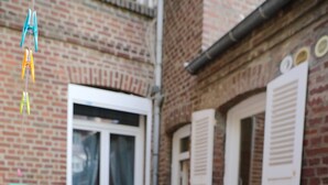 Een gebouw van baksteen met een grote tafel en stoelen, witte luiken en een raam met gordijnen.