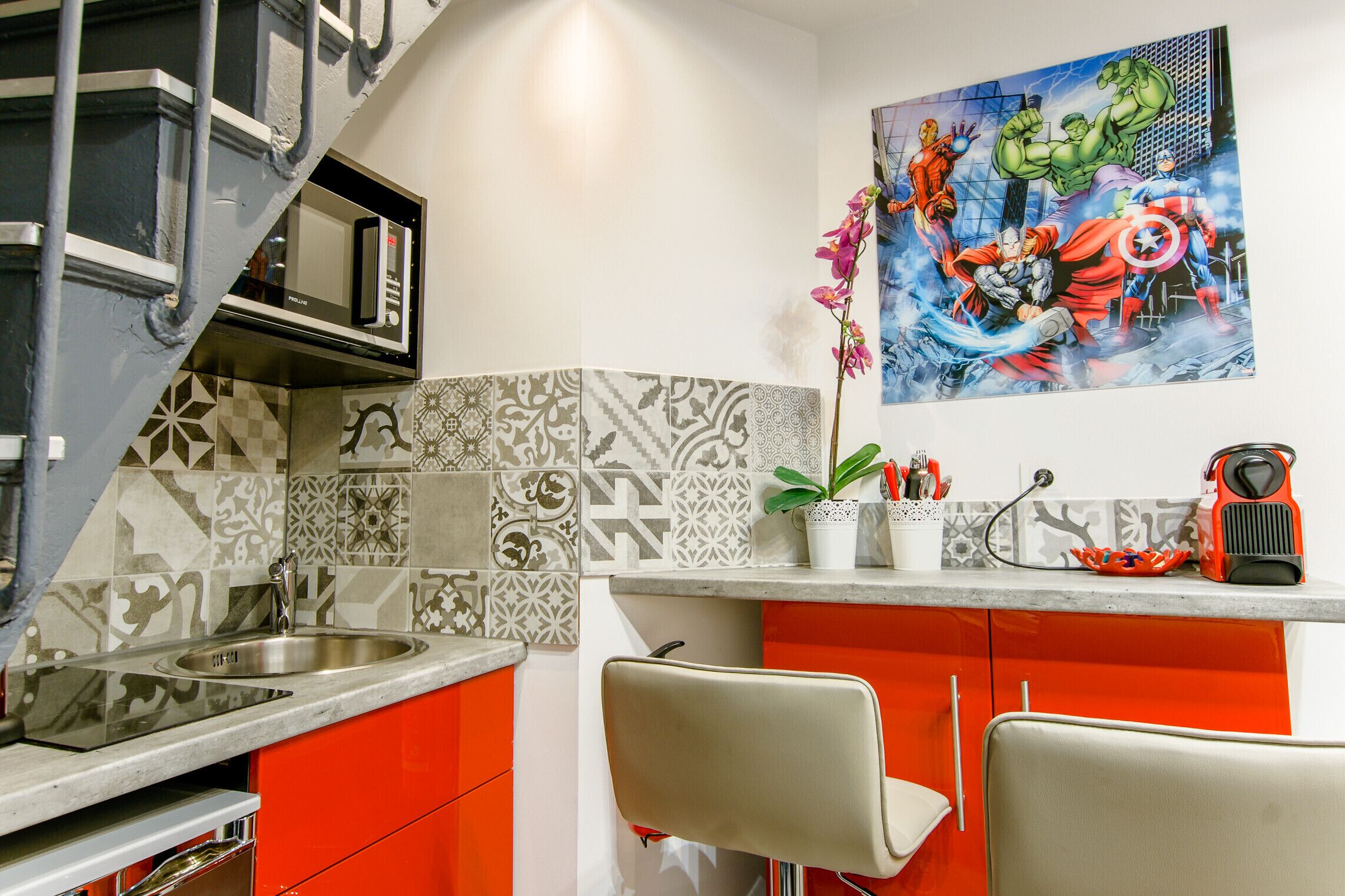 Una cocina con microondas, lavamanos y armarios color naranja. Se observa una silla y un adorno en la pared.