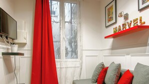 A living room with a red curtain, a grey sofa with red cushions, a mounted TV, and a shelf with colorful letters spelling "MARVEL".