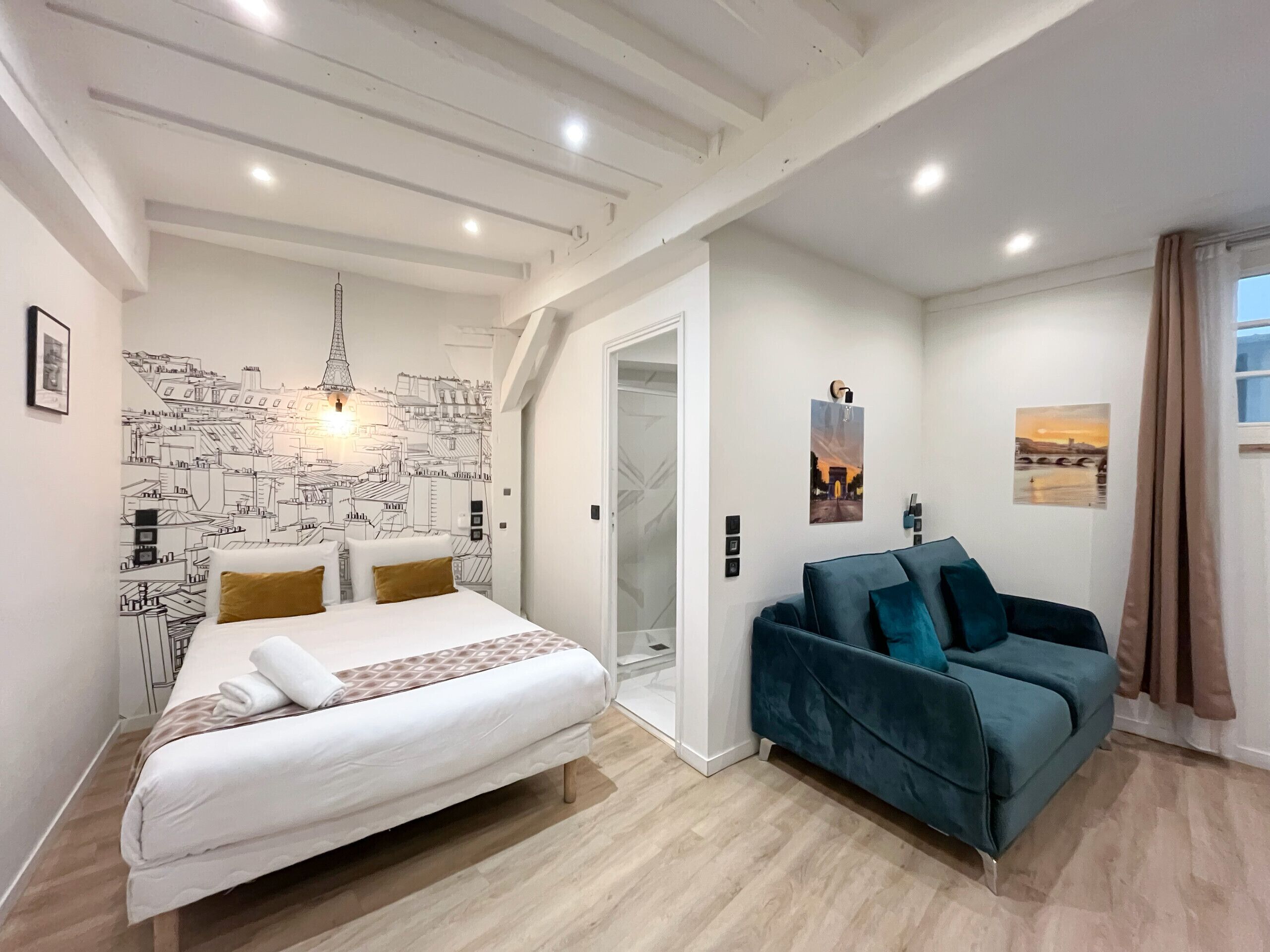 Une chambre moderne avec un lit, un canapé bleu, une fresque représentant un paysage urbain et une porte donnant sur une autre pièce.