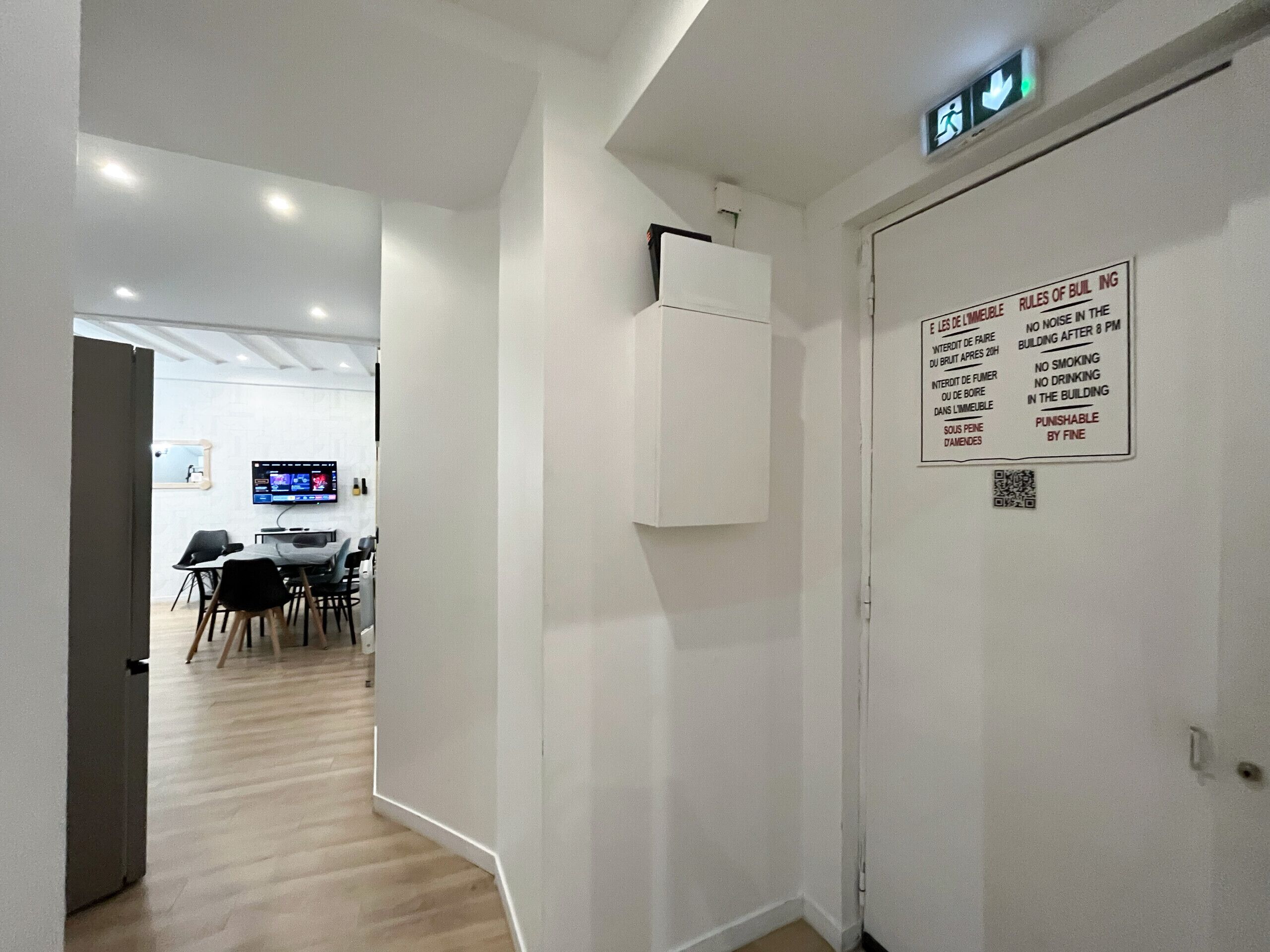 A hallway with a white door displaying rules of conduct, a conference room with a TV, and a wooden floor.