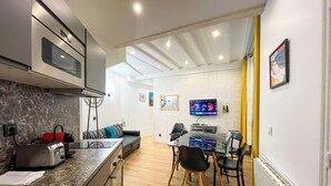 Una cocina moderna con horno y microondas integrados, un comedor con mesa y sillas de vidrio, y una sala de estar con sofá y televisor.