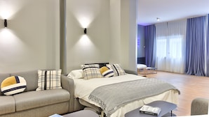 Et moderne hotelværelse med seng, sofa, lille bord og stol. To vægmonterede lamper og et stort vindue med gardiner.