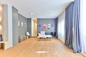 A spacious room with a wooden floor, a bed with a blue headboard, a desk with a chair, and a wall with framed pictures and a large painting.
