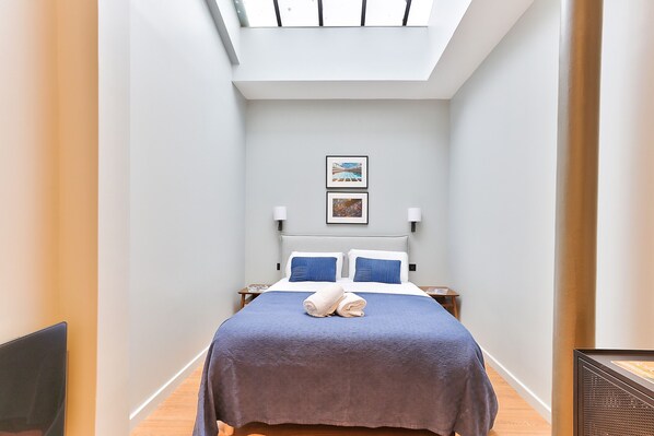 A narrow hotel room with a skylight, a bed with a blue bedspread, two pillows, and two wall-mounted lamps.
