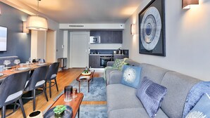 Una sala de estar moderna con un sofá gris, una mesa de comedor y sillas. Se ve una cocina al fondo, con un microondas y una estufa.