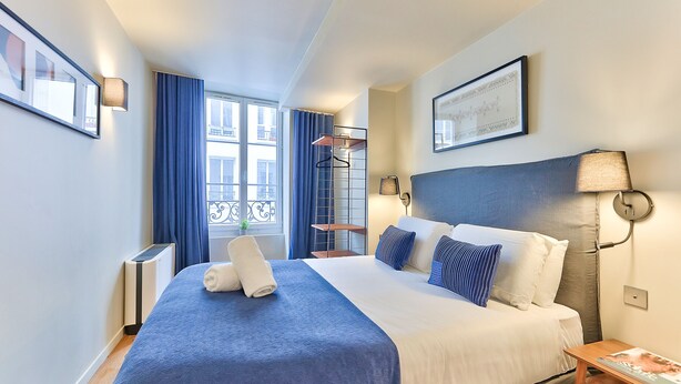 Una habitación de hotel con una cama grande, una mesita de noche, una ventana con cortinas, un estante en la pared y un cuadro enmarcado.