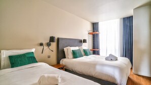 Une chambre d’hôtel avec deux lits, une petite table, un téléphone fixé au mur et une fenêtre avec des rideaux.
