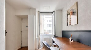 Ein Zimmer mit Holztisch, einem Stuhl, einer Heizung, einem Fenster mit Blick auf Gebäude und einem gerahmten Bild an der Wand.
