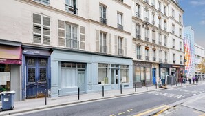 A street view with a row of buildings, including a blue storefront with a sign, a white building with shutters, and a colorful building with a unique pattern. - UrbanFlat 136 - Large Apartment in Paris Center (Paris)