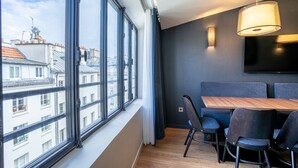 A modern room with large windows, a dining table with chairs, a flat-screen TV mounted on the wall, and a pendant light fixture.