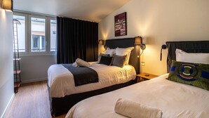 A hotel room with two beds, a window with curtains, a wall-mounted artwork, and a bedside table with a lamp.