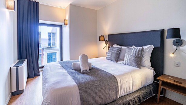 Ein modernes Hotelzimmer mit einem großen Bett, einem Nachttisch, einer Wandlampe und Blick auf die Stadt durch das Fenster.