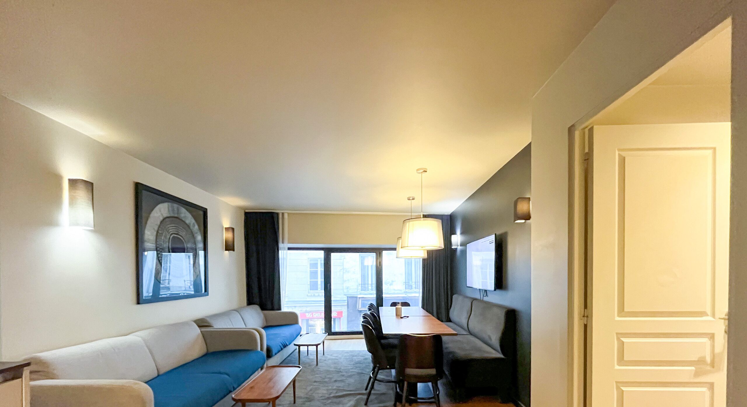 Un salon moderne avec un canapé, une table à manger et des chaises. Un téléviseur est fixé au mur et il y a une fenêtre avec des rideaux.