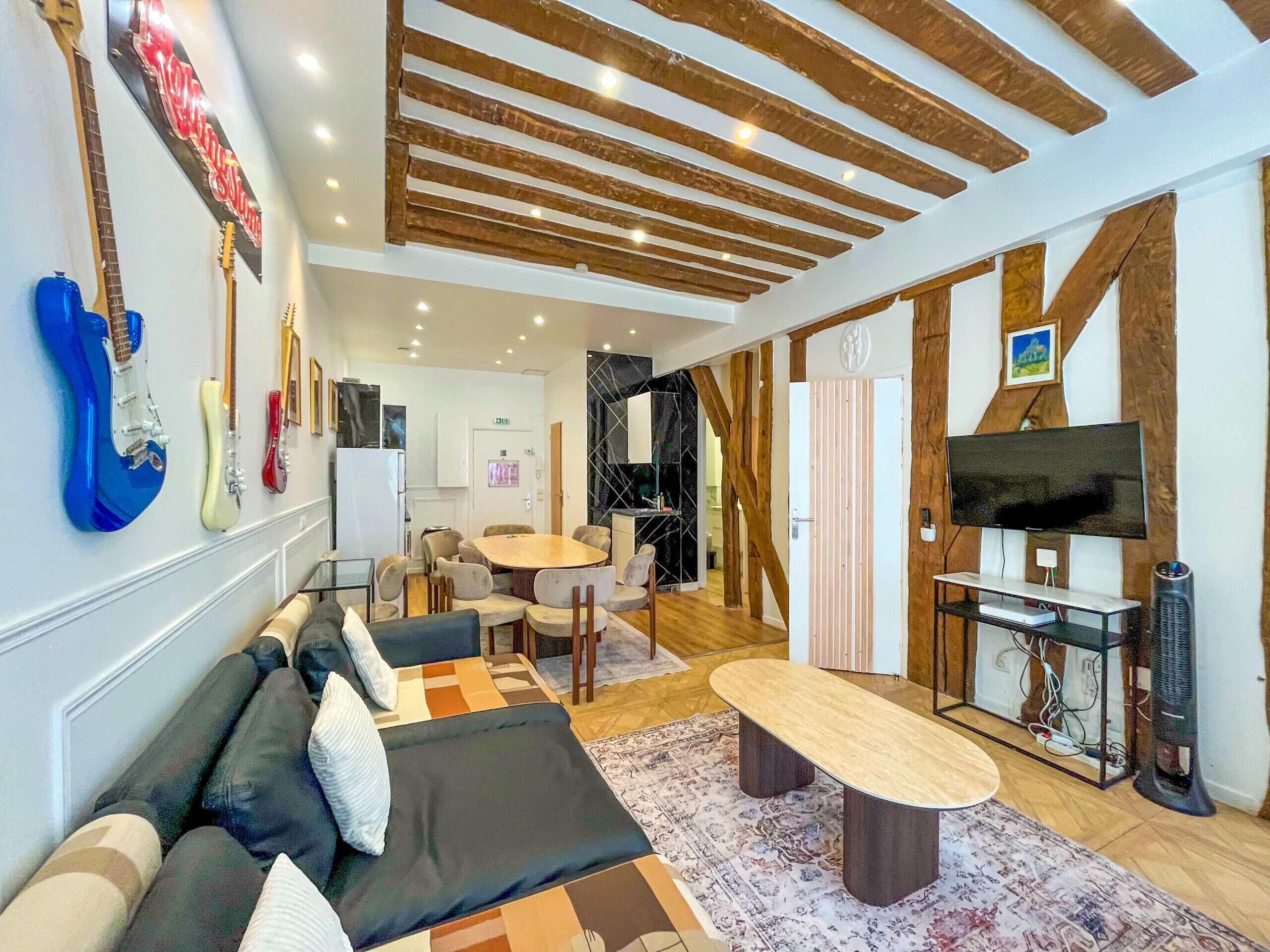A modern living room with a sofa, a coffee table, and a TV. There are guitars on the wall, a dining area, and a kitchen in the background.