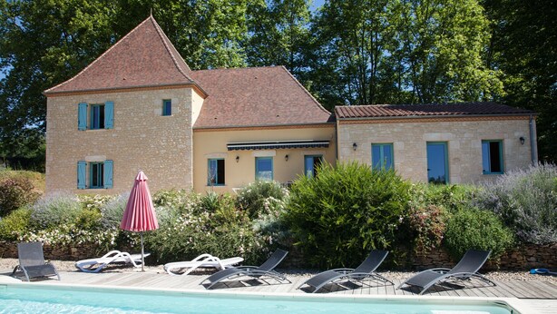 Un espace aménagé au bord de la piscine, avec des chaises longues et un parasol, entouré de bâtiments en pierre aux volets bleus.