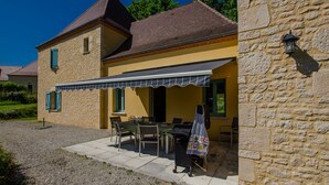 Un bâtiment en pierre avec une façade jaune, un patio couvert agrémenté de mobilier d’extérieur et une allée gravillonnée.