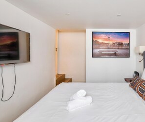 Una habitación de hotel con cama, televisión, un cuadro de un atardecer y una puerta que da a un pasillo.