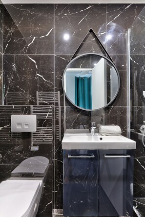 Ein modernes Badezimmer mit einer Wand aus schwarzem Marmor, einem runden Spiegel, einem weißen Waschbecken, einem Handtuchhalter und einer Dusche.