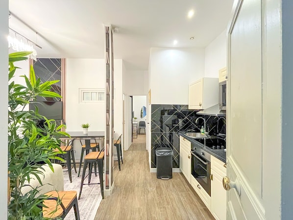 A modern kitchen with a black countertop, white cabinets, and a stainless steel sink. There is a dining area with a table and chairs, and a trash can in the corner.