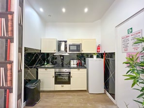 Een compacte keuken met witte kastjes, een magnetron, een oven en een koelkast. Er is een spoelbak met een kraan, een prullenbak en een brandblusser aan de muur bevestigd.