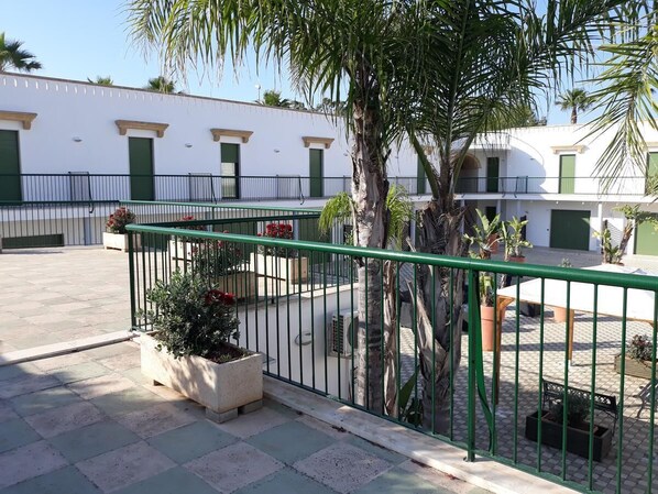 A courtyard with a tiled floor, a green metal fence, potted plants, and a white building with green doors and windows. - Cala Verde Residence - Two-room apartment for 4 people (Marina di Mancaversa)