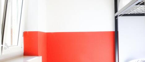 Una habitación pequeña y de colores vivos, con la mitad inferior roja y la superior blanca, una ventana, un radiador y un escritorio blanco.