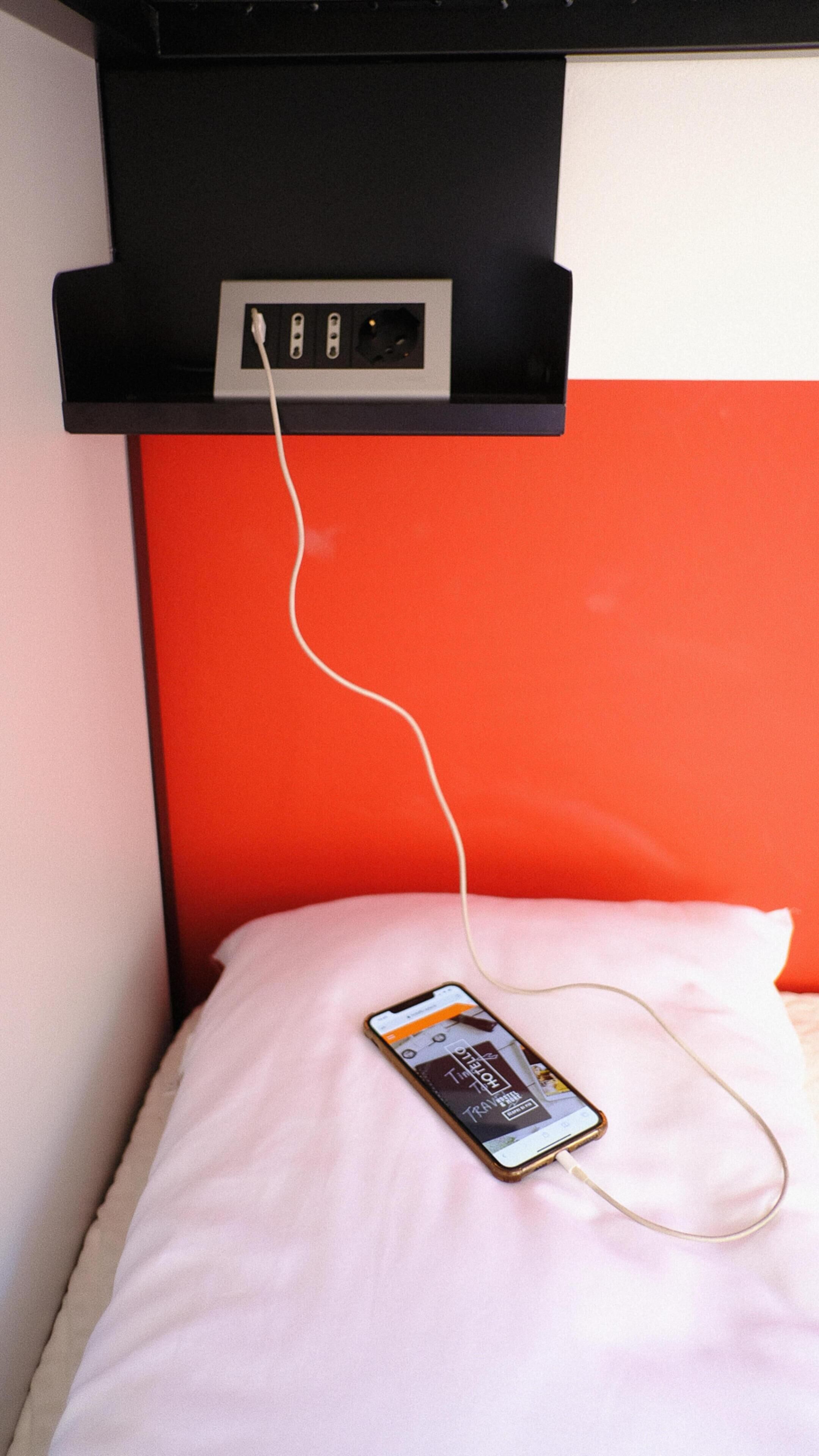 Un lit avec un oreiller rose et un téléphone intelligent posé dessus, branché à une prise électrique située sur une étagère noire au-dessus.