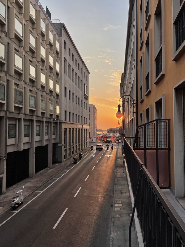 A city street at sunset with buildings on either side, a clear sky, and a few vehicles on the road.